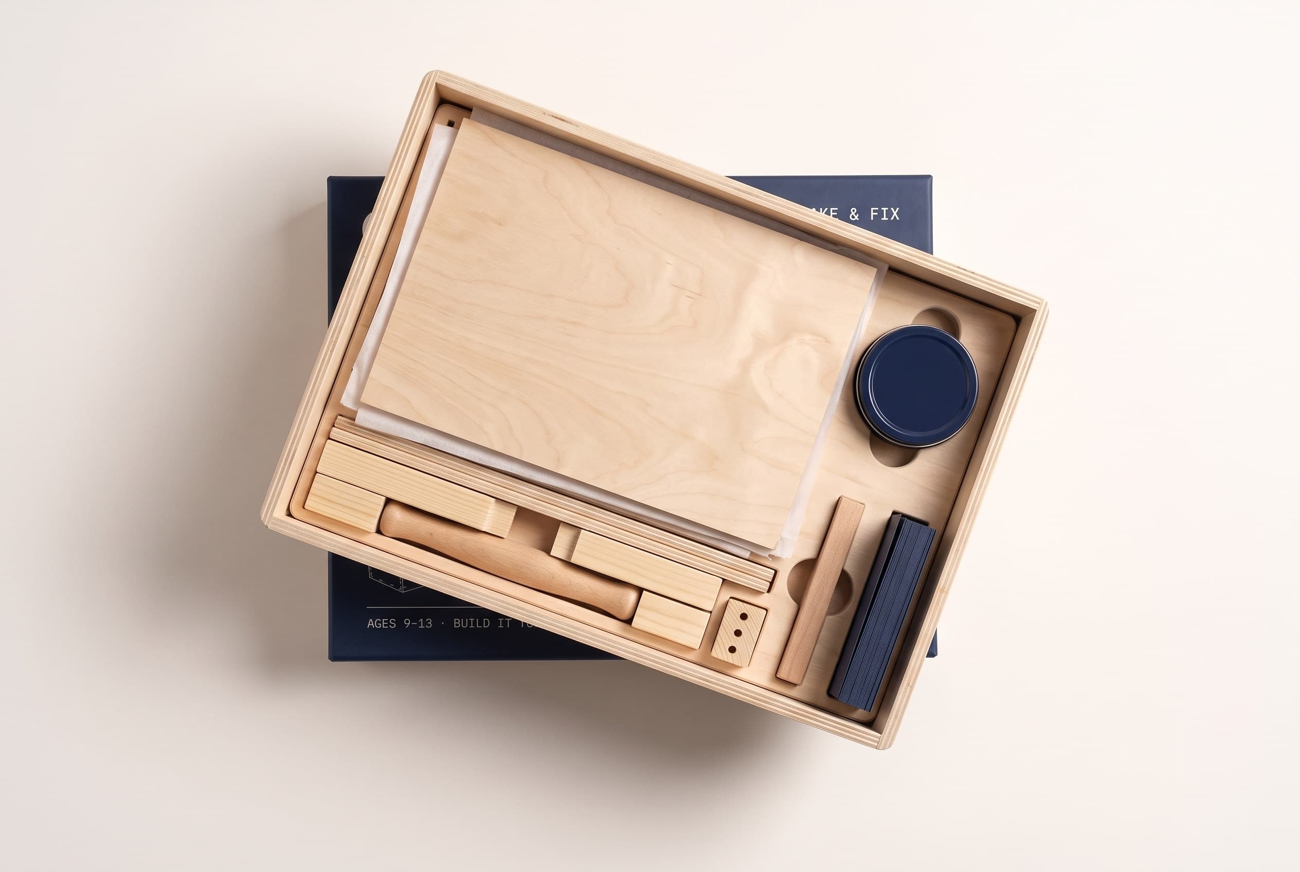 The open box seen from above: the practice board, hammer, screwdriver, ruler, safety glasses, sanding block, a tin of screws and nails and a stack of challenge cards, each seated in its own fitted recess, with the navy lid resting behind.